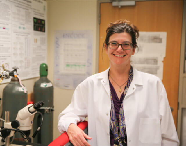 Woman with black hair wearing black glasses and white lab coat.