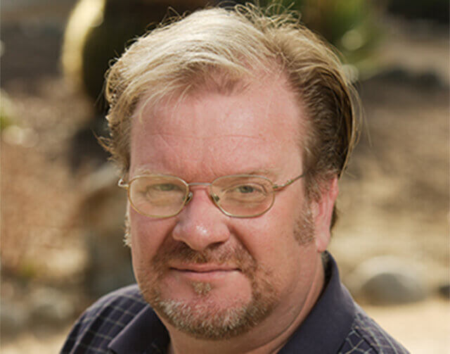 Man with short blonde hair, glasses, and a navy blue collared shirt.