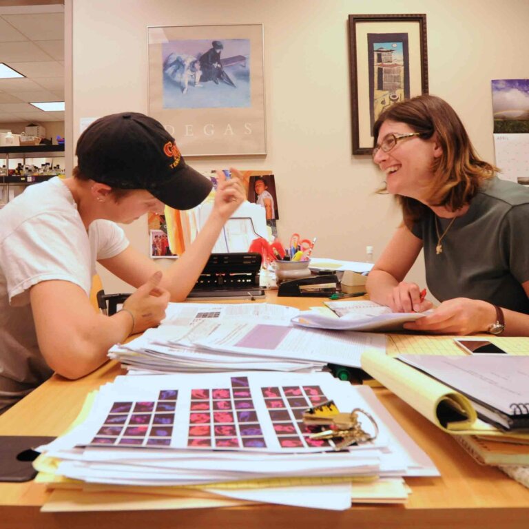 a student and professor sit opposite each other at a desk while looking over papers