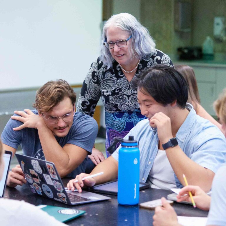 two students sit at a desk looking at a computer while a professor stands behind them explaining something.