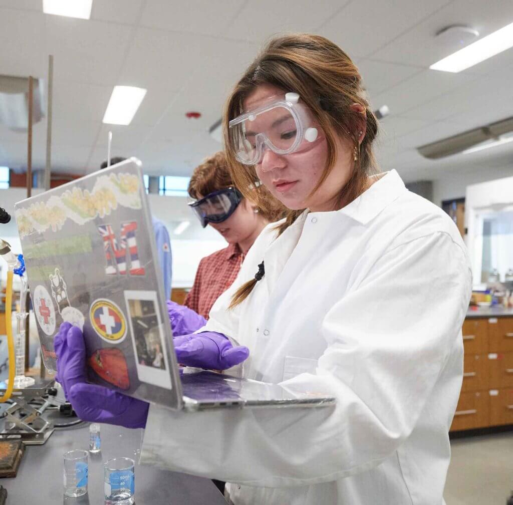a student in a white lab coat wearing google holds and looks at a laptop computer while standing in a lab.
