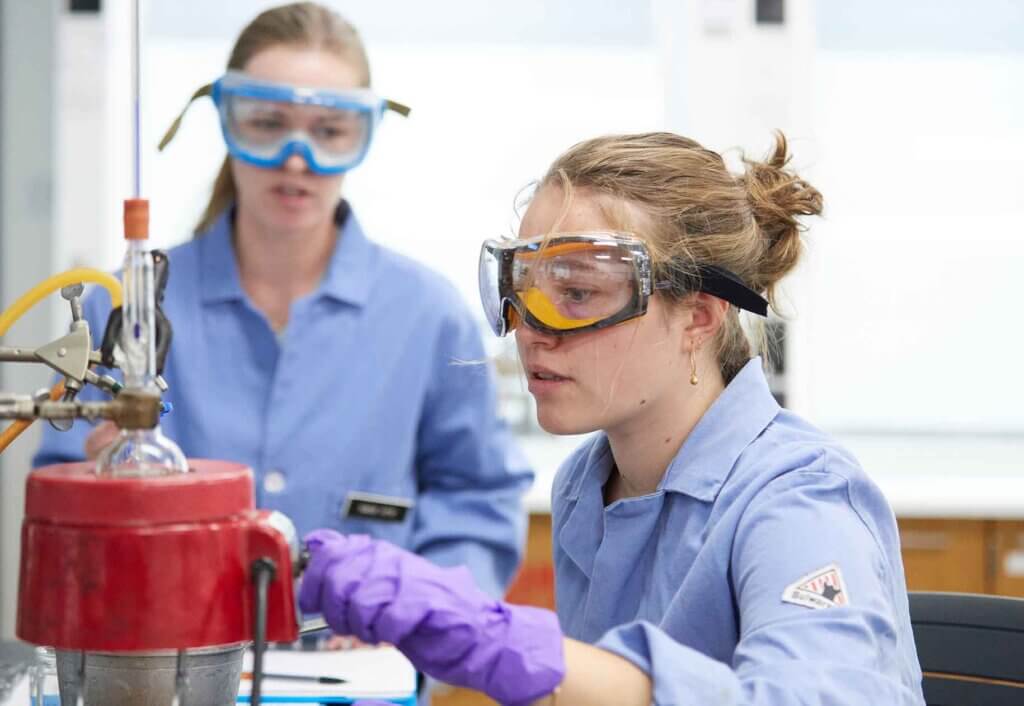 two students wearing blue lab coats conduct a chemistry experiment