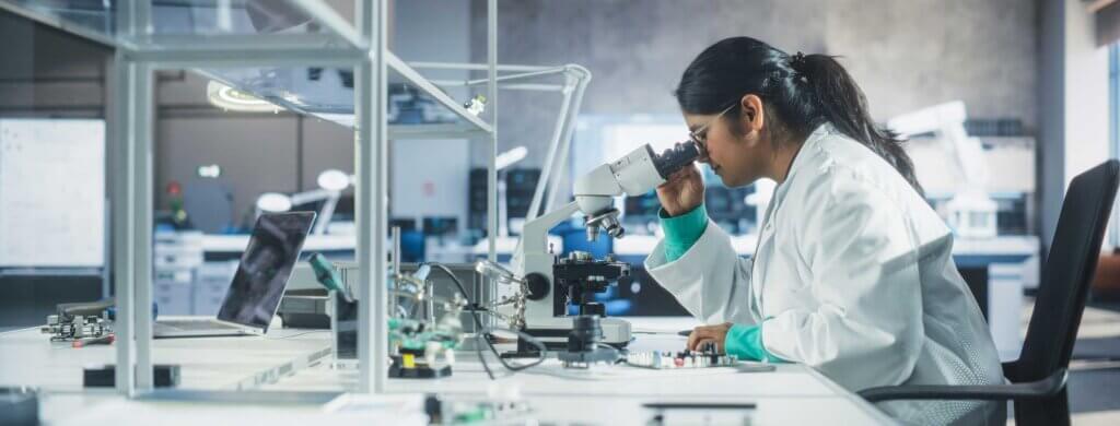 a woman with long brown hair, wearing a white lab coat, peers through a microscope in a lab.