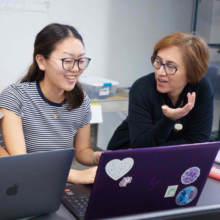 a student in a black and white striped tshirt and black glasses looks at a computer screen while a female professor explains to her