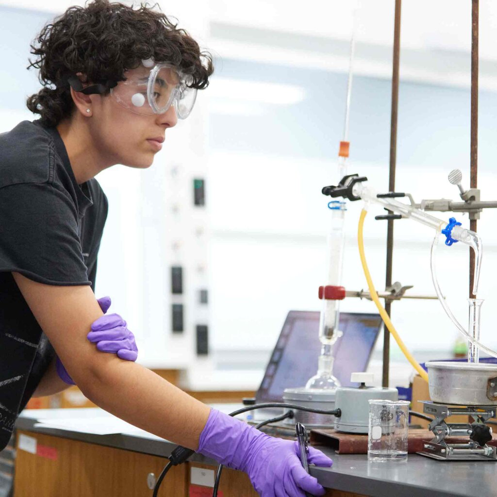 a student with short curly brown hair wears goggles and latex gloves while performing a chemistry experiment