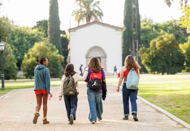 Four students, with their backs towards the viewer, walk towards a white, Spanish-style building surrounded by trees.