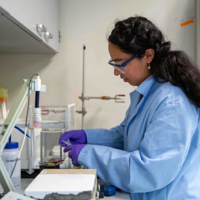 a student wearing a blue lab coat, gloves and protective eye gear works at a lab station