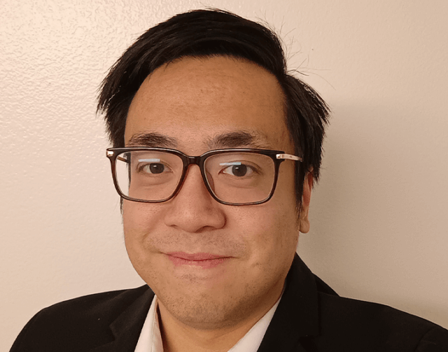 Man with short black hair in black and white suit and black glasses.
