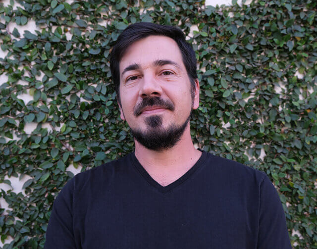 Man in black V-neck shirt standing and smiling in front of plant wall.