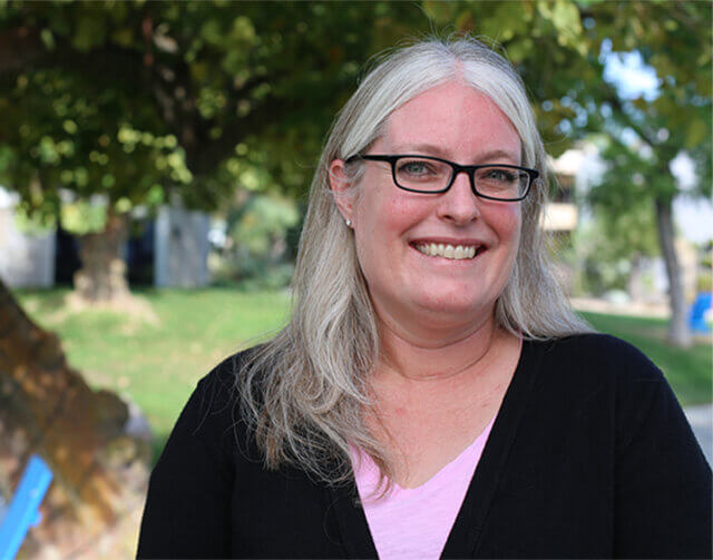 Woman with long white hair and black glasses wearing a pink and black blouse.