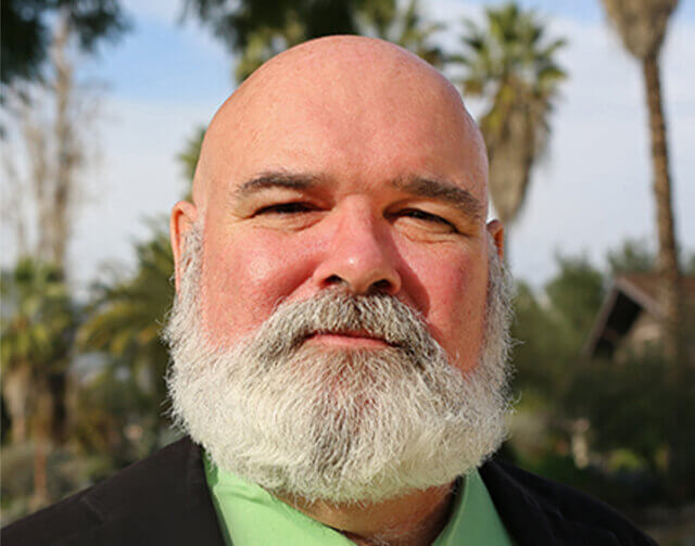 Man in black suit with green shirt in front of trees.