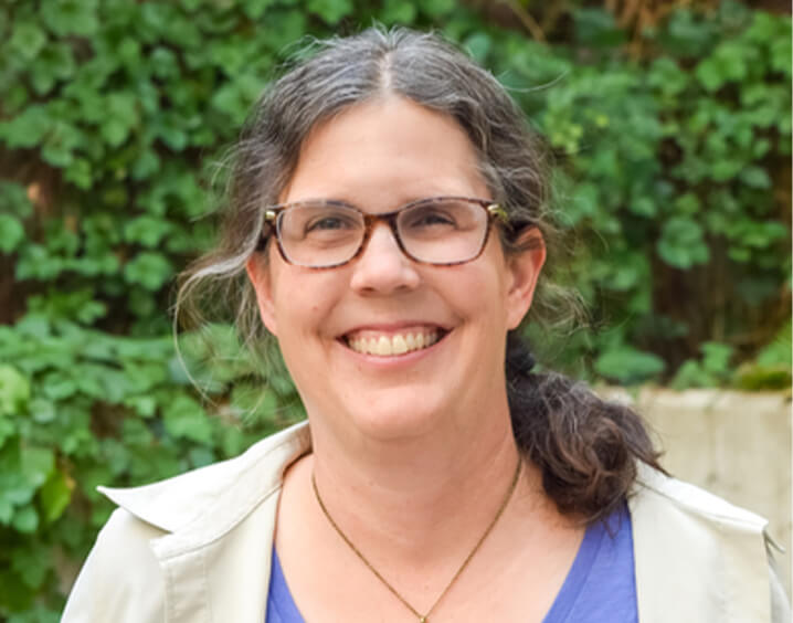 Woman in beige jacket and purple shirt with glasses.