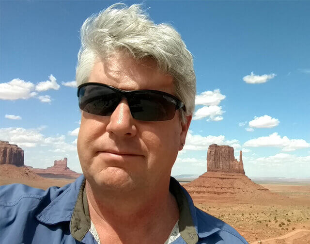 Man in blue shirt wearing black glasses standing in front of desert.