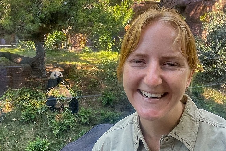 Jana Biedenweg stands in a panda enclosure while a panda behind her eats bamboo