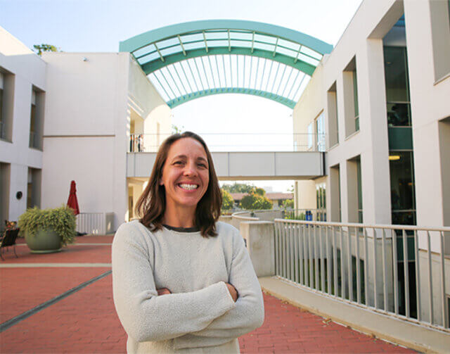 Woman with short light brown hair in white long sleeve sweater in front of buidling.