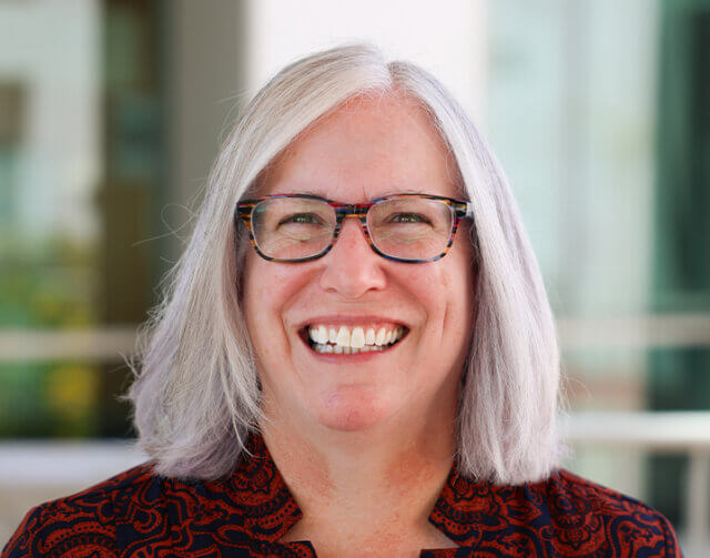 Woman with short white hair with black glasses and red-black shirt smiling.