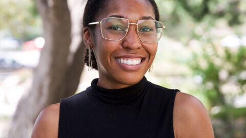 Woman in black shirt and clear glasses standing in front of tree smiling.