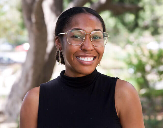 Woman in black shirt and clear glasses standing in front of tree smiling.