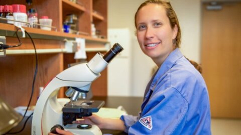 Professor Tessa Soloman-Lane wears a blue lab coat while sitting a microscope