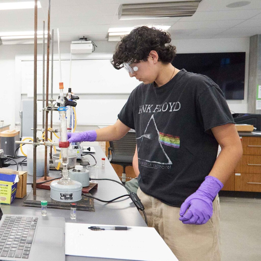 a student wearing goggle, gloves performs an experiment in a lab