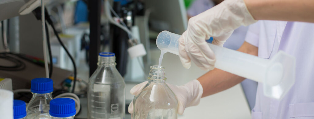 a pair of latex-gloved hands pours chemicals out of a beaker into a glass bottle