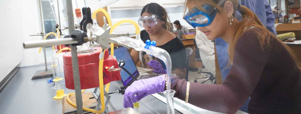 two students wearing goggles and latex gloves perform a chemistry experiment