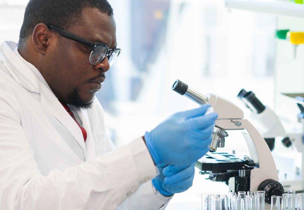 A chemistry student in a white lab coat and gloves works with vials next to a microscope