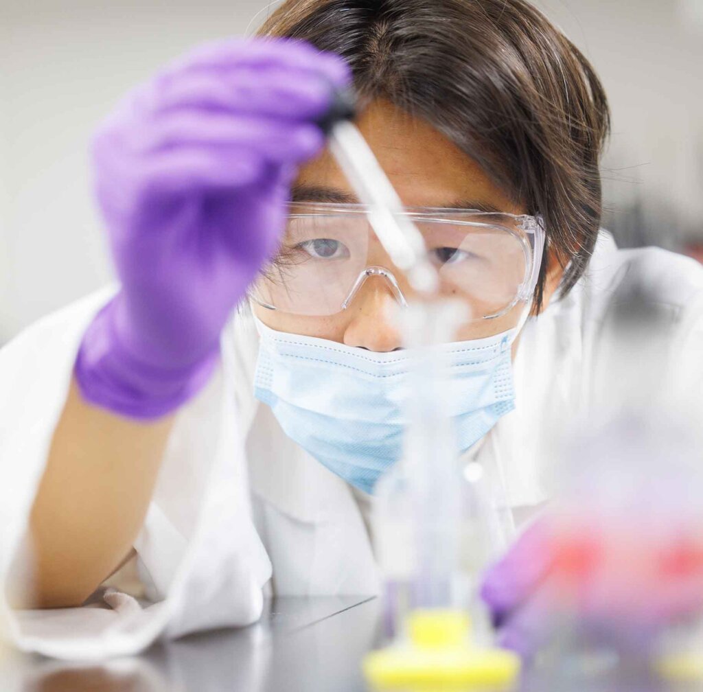 a student in goggles and gloves uses a dropper to add liquid to a vial