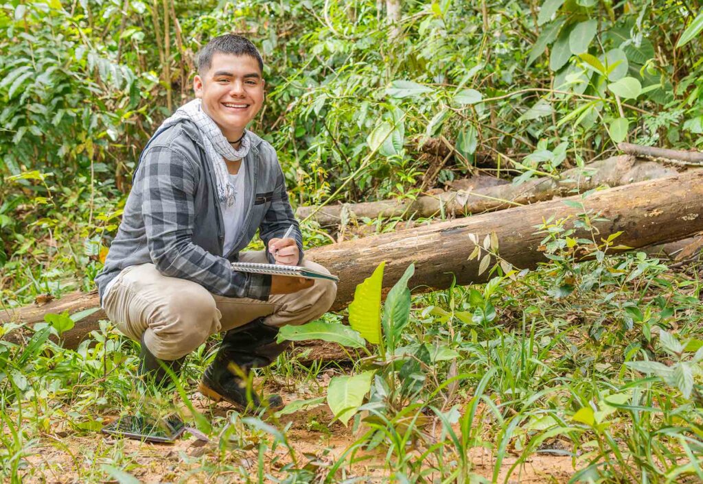 a student crouches down in a forest with a notebook in hand