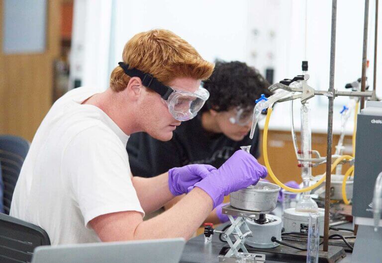 two students wearing goggles and gloves work with lab equipment