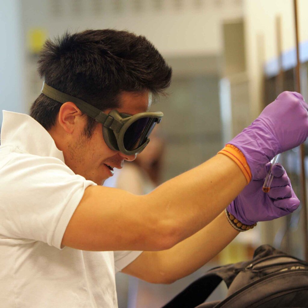 a male student with short black hair wears googles and latex gloves while workig with lab vials