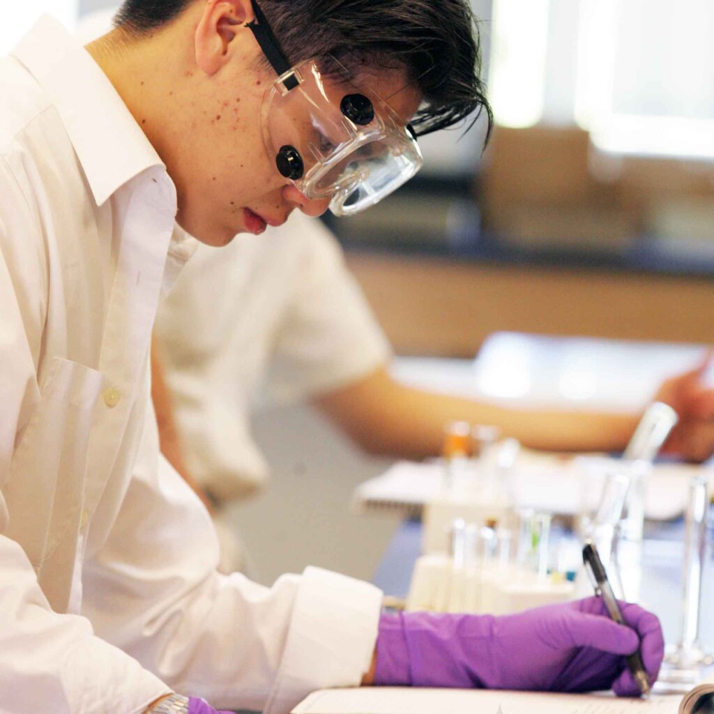 a male student wearing goggles and gloves takes notes in a lab
