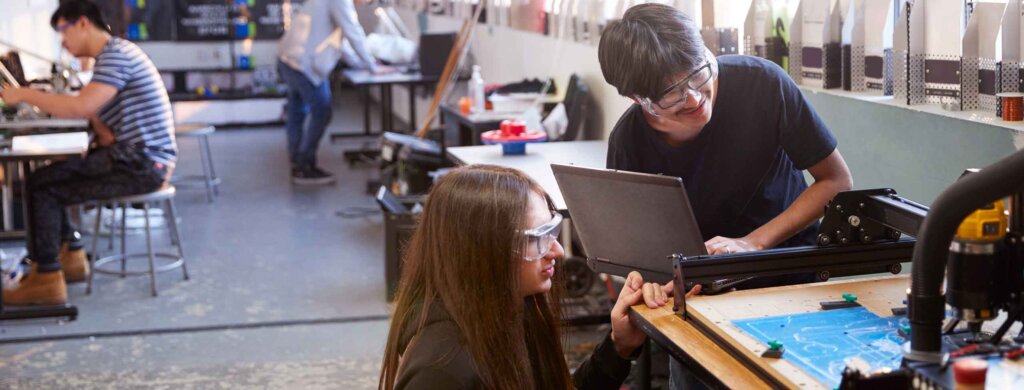 two students work at a 3d printer in a large room with other students