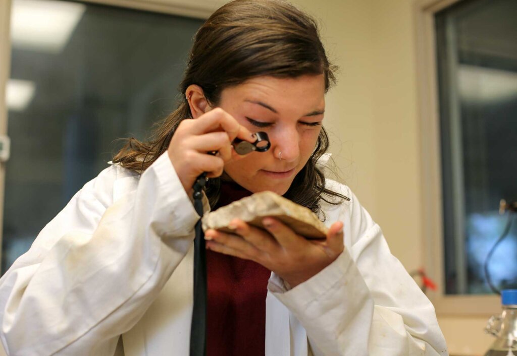 a woman wearing a white lab coat uses a magnifier to look at a specimen.