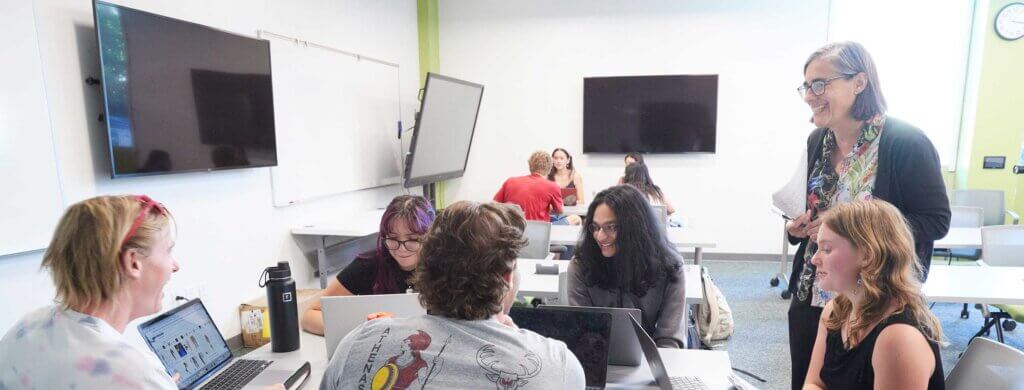 several student sit at a table while a professor speaks to them.