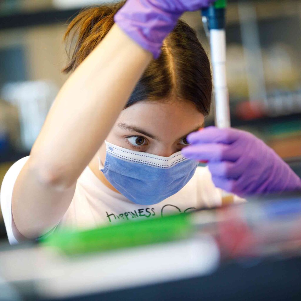 a student with a ponytail and surgical mask peers at a test tube