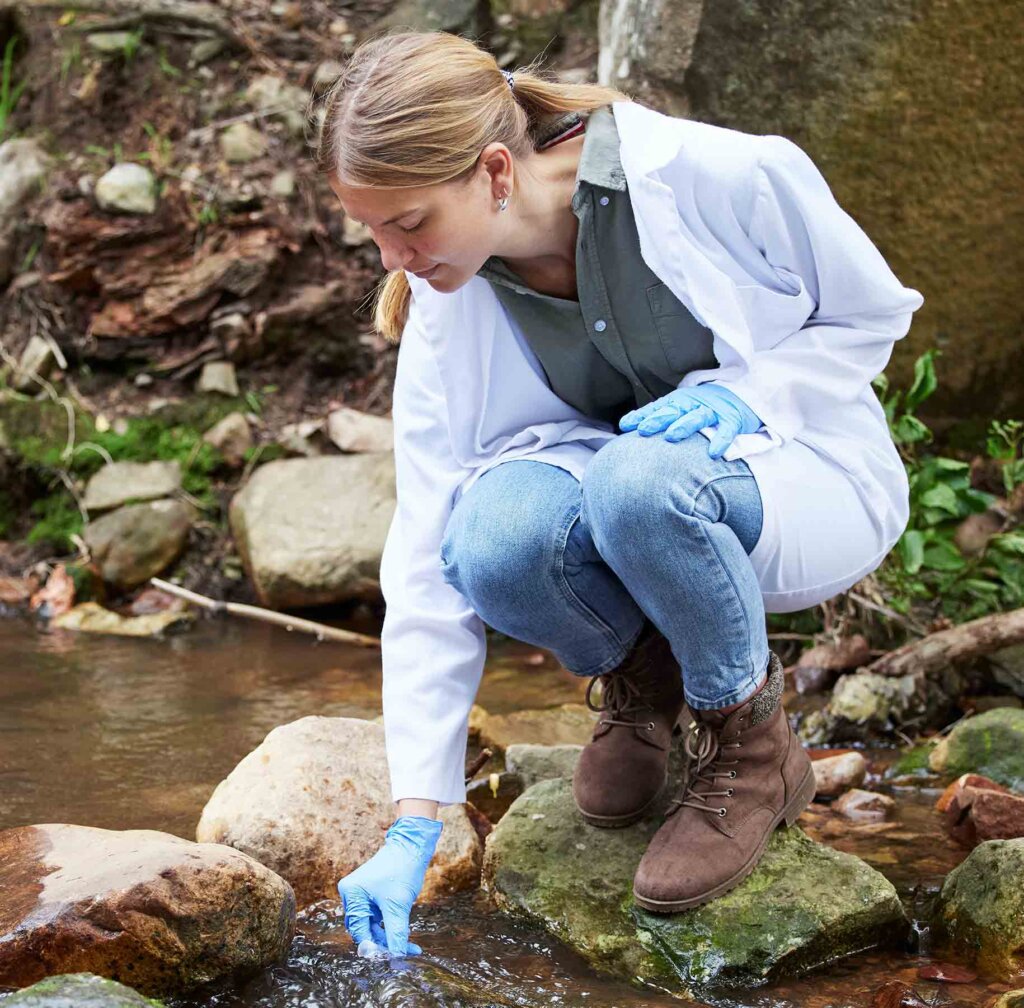 a woman wearing jeans and a lab coat fills a vial with pond water