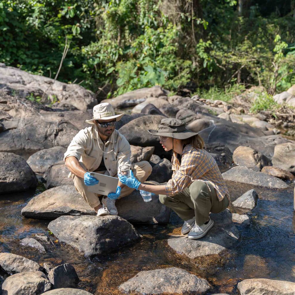 two students crouch on rocks while collecting water samples from a creek