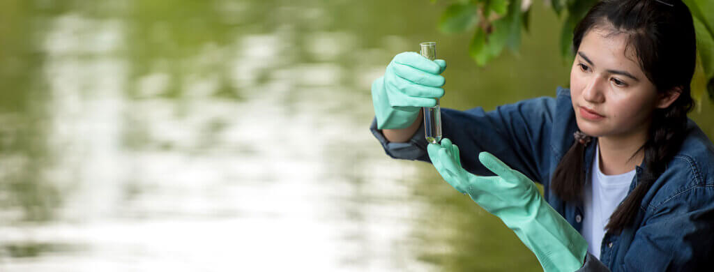 a young woman with long brown hair stands in front of a pond while holding a vial villed with murky water.