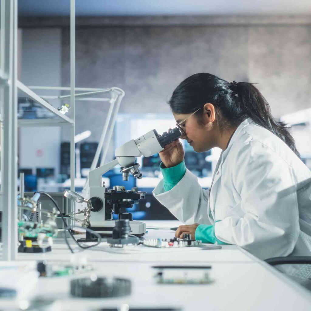 a woman with long brown hair in a ponytial and wearing a lab coat peers into a microscope in a lab.