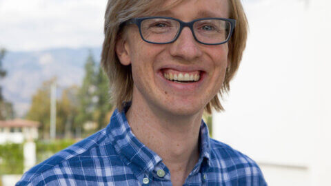 a man with ear-length blond hair wears black glasses and a blue and white plaid button up shirt
