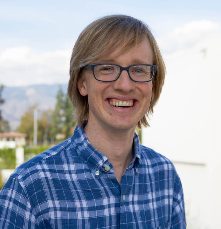 a man with ear-length blond hair wears black glasses and a blue and white plaid button up shirt