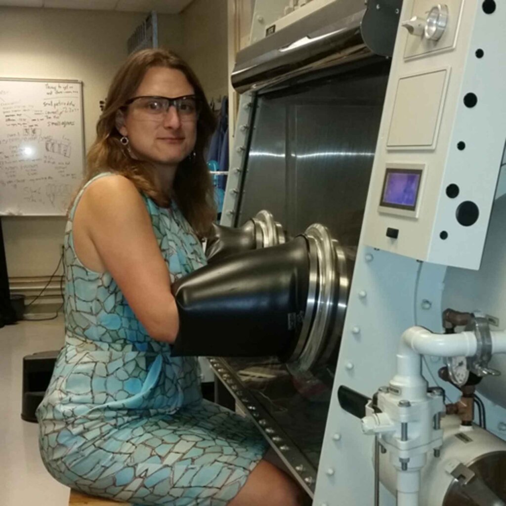 a woman with shoulder-length brown hair wearing a blue dress and glasses uses a glove box in a lab