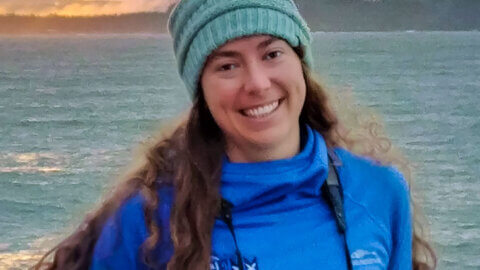 dawn barlow stands in front of water while wearing a pair of binoculars